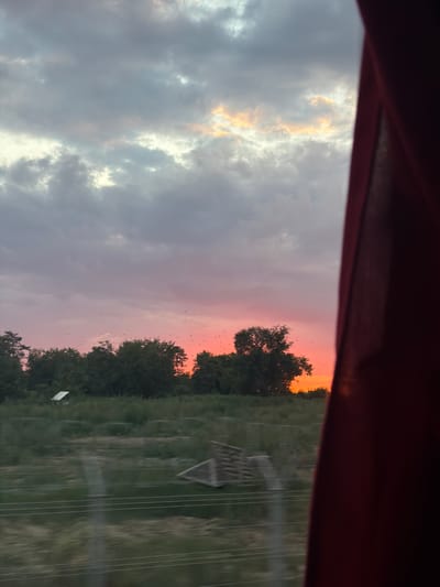 This is a photograph taken through a window, with a dark red curtain obscuring the right side of the frame. The image captures a sunset scene with a vibrant orange and pink sky. A line of trees forms the horizon, with a few scattered birds visible against the colorful sky. In the foreground, there is a blurry grassy field with what appears to be a small structure and some wooden debris. Power lines run horizontally across the lower portion of the image, adding a sense of depth and perspective. The overall impression is of a serene, natural landscape bathed in the warm glow of the setting sun. The location is identified as Unknown, Romania.