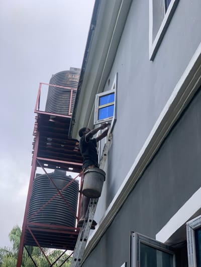The scene, located in Amaeshi, Nigeria, shows an outdoor view of a grey-painted building facade under an overcast sky. A dark-skinned male, wearing a dark t-shirt and dark trousers, stands on a silver metal ladder. He is positioned against the building at the level of a second-story window, which has a white frame and a blue-tinted pane, and is actively working on its exterior frame with both hands. A grey metal bucket is suspended from the ladder below his right foot. To the left of the building, a red metal scaffolding structure supports two large, ribbed, black plastic water tanks; the upper tank bears a partial "HART Industrial Ltd" label. Green foliage is visible in the lower-left background, and a partially open window with "aluminium global aluminium profe" visible on its frame is present in the lower right foreground.