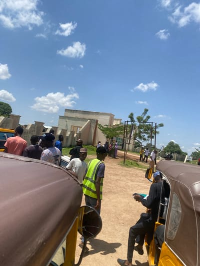 In Yali, Nigeria, a group of people are gathered outdoors under a bright blue sky with scattered white clouds. The scene appears to be near a building with light-colored, somewhat weathered walls and decorative concrete elements. In the foreground, the tops of two brown auto-rickshaws, also known as "keke napep," are visible.
Several individuals are standing around, some appearing to be waiting or observing. One person in the mid-ground wears a bright yellow-green reflective vest. Another man, further to the right and seated in an auto-rickshaw, is dressed in black and appears to be holding a book or tablet. The environment is dusty or sandy, with some patches of green grass and trees in the background. The overall impression is of a public space where people are congregating.