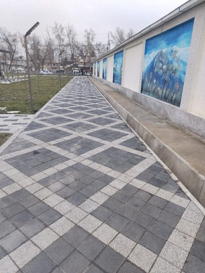 The image displays a pedestrian pathway in Chighirik, Uzbekistan, observed under an overcast sky. The pathway is constructed from interlocked concrete pavers arranged in a repeating diamond pattern using alternating dark gray and light gray blocks, extending into the background. To the right of the pathway, a concrete drainage channel runs parallel to a light-colored wall. This wall is adorned with multiple uniformly-spaced rectangular murals, each depicting a blue-skied mountainous landscape with trees. To the left of the pathway lies a strip of grassy ground with sparse vegetation, along which several dark-colored streetlights are positioned. In the distant background on the left, a street is visible with several white vehicles, likely vans or cars, parked near multi-story buildings and numerous bare deciduous trees. No individuals are present within the frame.