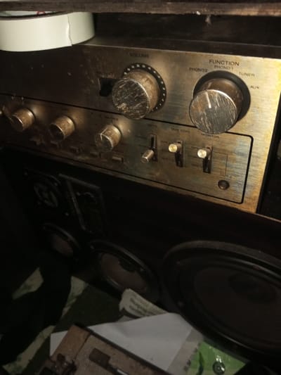 The image shows a dusty, vintage audio amplifier and speaker system. The amplifier's front panel features knobs for volume and function selection (phono, tuner, aux), as well as switches for mode, tape copy, and monitor. A series of smaller knobs sits below the larger ones. The speaker cabinet is positioned beneath the amplifier. Both units appear to be aged and in need of cleaning.