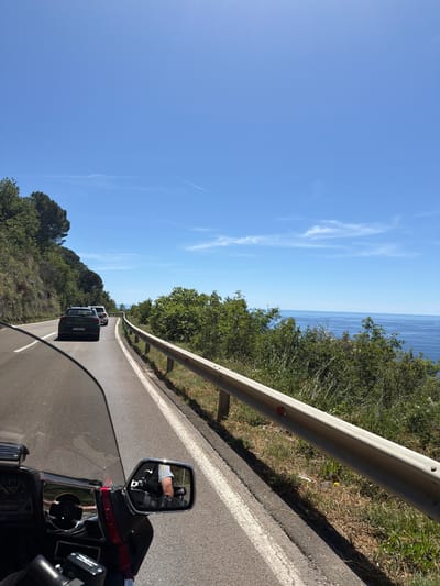 This image depicts a scenic view from the perspective of a motorcycle rider on a sunny day. The road curves to the right, with a steep, tree-covered hillside on the left and a guardrail separating the road from a drop-off to the ocean on the right. The ocean is visible in the distance, a deep blue under a clear sky with a few wispy clouds and contrails. Two cars are driving ahead on the road, with a darker SUV in the foreground followed by a lighter vehicle. The motorcycle's windshield and dashboard are visible in the lower left corner, with a side mirror reflecting the rider wearing a black vest with "bm" logo and black gloves. The environment suggests a coastal highway, likely in a Mediterranean or similar climate, consistent with the provided location of Gornji Katun, Montenegro, which is known for its coastal roads and natural beauty.