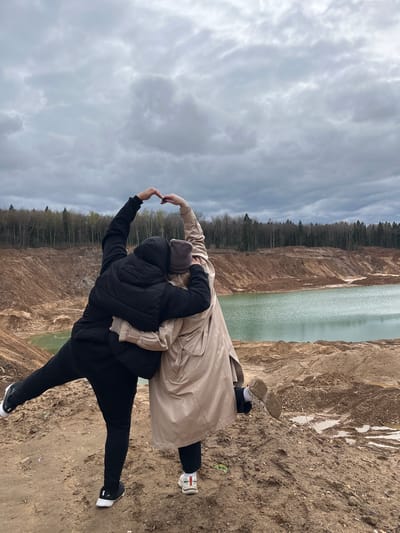 Two people are standing on a sandy slope in Ivlevo, Russia, appearing to form a heart shape with their hands. The person in the foreground is wearing a black puffer jacket and black pants with black sneakers, while the person behind is wearing a beige trench coat, black pants, and white sneakers. A quarry filled with turquoise water is in the background, along with a wooded area and a cloudy sky.
