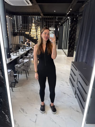 A woman with long brown hair stands in a salon or spa, taking a mirror selfie. She is wearing a black, sleeveless, zippered jumpsuit and black Crocs. The background shows a modern interior with marble flooring, black curtains, and a contemporary staircase with ornate lighting. To her right, there is a gray dresser with multiple drawers. In the reflection of the mirror, other salon chairs and workstations are visible. The woman is holding a white smartphone in her right hand. The location is identified as Укромновское сельское поселение, Ukraine.
