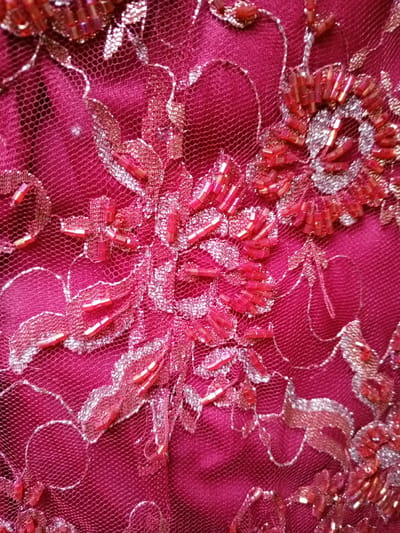 This image is a close-up, detailed shot of an intricately embellished textile, likely a piece of fabric used for clothing or decoration. The background is a deep magenta or dark pink, composed of a fine, sheer mesh or tulle fabric, possibly layered over a solid lining of the same color.
The textile is adorned with elaborate embroidery, featuring floral or organic patterns. These patterns are created using two primary decorative elements: numerous small, cylindrical (bugle) beads and metallic thread. The beads are predominantly red and crimson, with some appearing iridescent, and are densely arranged to form various parts of the stylized floral designs, such as petals and central motifs. Complementing the beads, metallic thread, which appears silver or light gold, is used to outline the embroidered patterns and fill in other sections of the designs, adding a shimmering contrast against the deep pink fabric.
The image highlights the rich textures and reflective qualities of the materials. No people are visible, and no specific activity is taking place. The focus is entirely on the craftsmanship and detail of the textile. There are no discernible time-of-day or weather indicators, nor are there any visible location cues beyond the textile itself. No text is visible in the image.