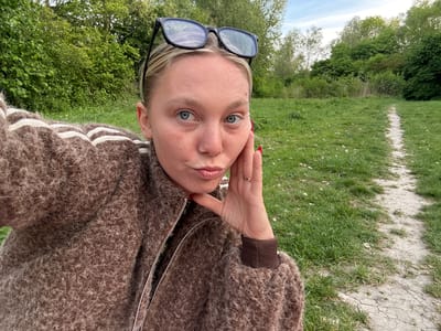 A person with blonde hair and blue eyes is photographed from a close-up, upside-down perspective. They are wearing black-rimmed glasses and a brown, textured jacket with a zipper. The background consists of green grass, trees, and a dirt path, suggesting an outdoor environment. The location is not conclusively identifiable as Marquette-lez-Lille, France, based solely on the image.