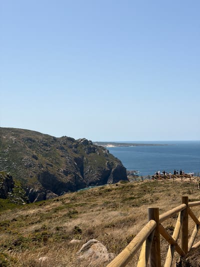 The image shows a view of the Atlantic coast in Colares, Portugal. In the foreground, a rustic wooden fence runs diagonally across the frame, with dry, golden grass and scrub covering the hilly terrain. In the midground, a group of approximately 10 to 15 people are standing on a wooden viewing platform, looking out at the sea. Beyond the platform, a steep, rocky cliff face descends to the water. The sea is a deep blue, meeting a distant, sandy coastline under a clear, pale blue sky. The landscape is characterized by its rugged, natural beauty.