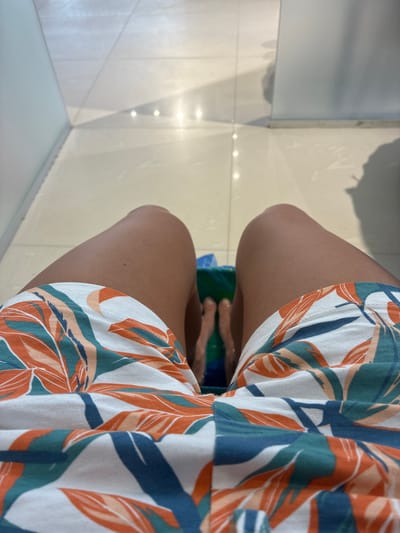 A first-person perspective shot shows a person's legs, clad in white shorts with an orange, green, and blue leaf pattern, resting in what appears to be a nail salon. The person's feet are submerged in a green foot bath, visible just below the hem of the shorts. The floor is tiled in a light, neutral color, and the overall environment suggests a clean, well-lit indoor space, possibly in Khashuri, Georgia, a city known for its diverse landscape and growing tourist industry.