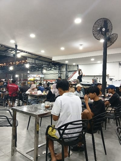 The image depicts an indoor food court or large casual dining area located in the city of Seumadam, Indonesia. The floor is light grey, and the ceiling transitions from a flat white surface with embedded spotlights in the foreground to an exposed industrial-style corrugated metal roof with suspended lighting in the background.

Approximately twenty-five individuals are visible, primarily adult males, seated at various tables. In the immediate foreground, two males occupy dark plastic chairs at a rectangular metal table with a reflective surface. The male with his back to the camera wears a white short-sleeved polo shirt and yellow shorts; a glass containing a dark beverage is on the table in front of him. The male seated to his right wears a black short-sleeved t-shirt and dark shorts, with his hand near his mouth. Other patrons include individuals in red and black t-shirts and a female wearing a light-colored head covering (hijab).

In the mid-ground, a person in a dark shirt is standing on a red and white step ladder or platform, holding an object above their head, possibly engaged in maintenance or setup. Two large, black, industrial-style oscillating fans are mounted on a tall black pole positioned on the right side of the frame, extending towards the ceiling. Multiple food stalls with partial Indonesian signage and various equipment are visible in the background. The scene suggests a busy social and dining environment.