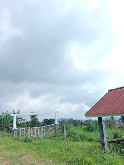 The image captures an overcast outdoor scene in the city of Nam Sa Lat, Myanmar. The upper two-thirds of the frame are filled with a bright, cloudy sky, characterized by diffuse grey and white clouds. The lower third consists of uneven green grassy terrain with varying heights of vegetation. In the midground, slightly to the left, a white-painted rectangular archway stands, comprising two vertical pillars with stone bases and a horizontal connecting beam. Immediately in front of this arch, a partially assembled or dismantled light-colored metal fence or railing with a criss-cross design is present. Banana trees are visible to the far left of the archway. To the right of the frame, a segment of a small, open-sided shelter is visible, featuring a weathered red corrugated metal roof that slopes downwards to the right, supported by a dark-colored post. A crude wooden bench or platform is situated beneath this shelter. The background reveals dense green foliage, numerous trees, and distant hills or mountains, with faint outlines of scattered structures visible on the far horizon. No individuals are depicted, and no actions or interactions are occurring.