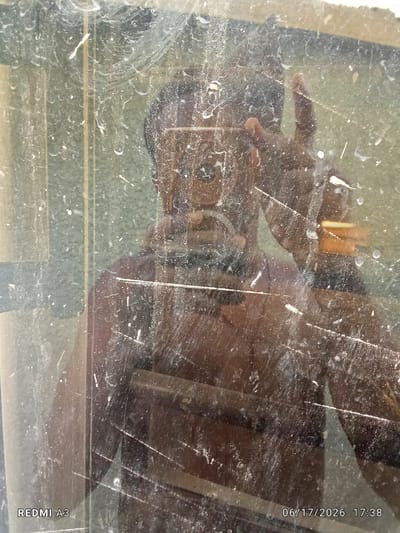 The image shows a reflection of a person in a dirty mirror. The person appears to be a young man, possibly of African descent, with dark skin and short, dark hair. He is wearing a dark, sleeveless shirt. He is holding up his right hand, with his index and middle fingers extended, as if giving a peace sign. The reflection is distorted and obscured by numerous smudges, dust particles, and streaks on the mirror's surface, giving it a grimy appearance.
The background reflected in the mirror is blurred and indistinct. It appears to be an indoor setting, possibly a room with a green-colored wall or surface behind the person. There are also horizontal lines visible in the reflection, which could be the edges of the mirror frame or other objects in the room. The lighting suggests it might be daytime, but the overall quality of the reflection makes it difficult to ascertain specifics about the environment.
The image is a selfie, taken by the person himself. The timestamp in the bottom right corner indicates the date and time as "06/17/2026 17:38". The text "REDMI A3" is visible in the bottom left corner, suggesting the phone model used to capture the image. The overall mood is informal, possibly a casual self-portrait taken in a personal space. The smudges on the mirror are a prominent feature, significantly impacting the clarity and aesthetic of the photograph.