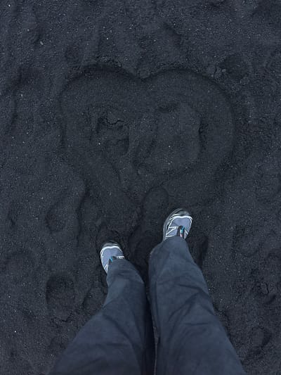 A high-angle shot shows a pair of legs wearing grey hiking shoes and dark grey pants standing on a black sand beach, likely in or near Vik, Iceland. A crude heart shape has been drawn in the sand to the left of the legs. The sand is coarse and dark, typical of volcanic beaches.