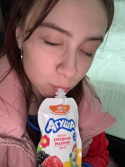 A woman is lying down and appears to be drinking from a white and blue fruit puree pouch labeled "АГУША". The text on the pouch indicates it contains apple, blackberry, and raspberry puree. The woman has closed eyes, brown hair, and is wearing a light pink jacket. The location is Lunezhki, Russia.
