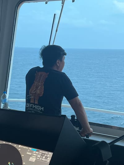 A person wearing a black t-shirt with a graphic of a character and the words "OFFHIGH" is standing on a boat and looking out at the ocean through a window. The person's back is to the camera, and they are leaning on a railing. A bottle of water is on the windowsill next to them. In the foreground, a large black screen displays radar information. The scene appears to be on a ship at sea, under a cloudy sky.