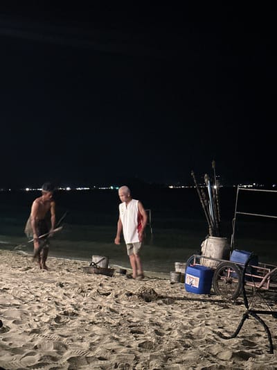 At Baan Klong Prao, Thailand, the scene features two people on a sandy beach at night. One man, wearing a white shirt and shorts, stands upright. The other, shirtless and facing away, appears to be working with long sticks. Behind them is a cart carrying supplies, including blue containers and what appears to be fishing equipment. The background is a dark sky with a few distant lights that appear to be city lights. The beach has distinct footprints and patterns in the sand.