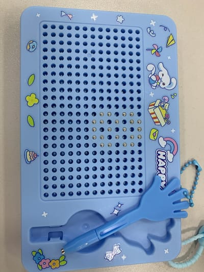 An overhead, eye-level medium shot captures a light blue magnetic drawing board with a grid of small, circular indentations, presumably for magnetic beads. The board is adorned with various cartoonish stickers, including a smiling bunny, a stack of presents, a rainbow, and colorful flowers. A blue stylus, shaped like a hand with outstretched fingers, rests in a molded holder on the lower portion of the board. A beaded blue lanyard is attached to the upper right corner of the board. The board itself is placed on a light-colored, textured surface, likely a table. The scene is devoid of any contextual clues to determine its specific location within Urtaaul, Uzbekistan. There are no people or actions depicted in the image.