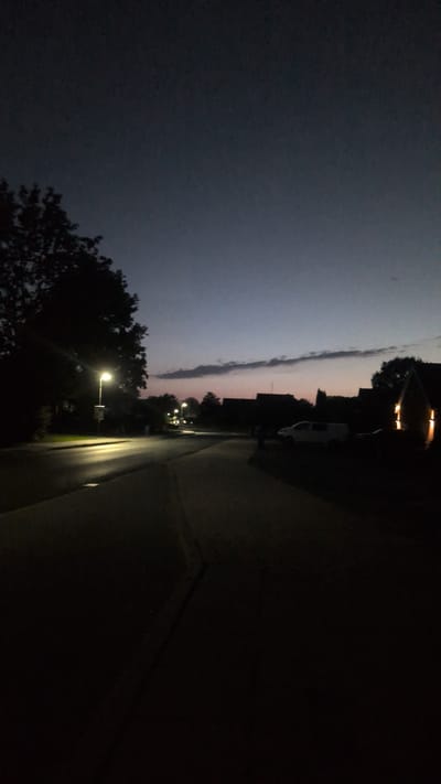 The image depicts a street scene at dusk in Hetlingen, Germany. A road curves into the distance, illuminated by a streetlamp on the left side, casting a bright pool of light on the asphalt. Trees line the left side of the road, silhouetted against the darkening sky. On the right, houses are visible, with some lights on, creating warm glows. A white van is parked on the right side of the road. The sky is a deep blue, transitioning to lighter shades near the horizon where a faint, elongated cloud formation is present. The overall atmosphere is quiet and serene, characteristic of a residential area at the end of the day.