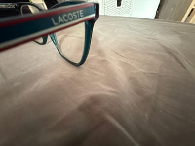The image shows a close-up of Lacoste eyeglasses resting on a wrinkled, dark gray fabric surface. The glasses have a teal frame with a red and white stripe detail on the temple. The background is blurry, but a white woven object and a beige vertical surface can be discerned. The scene's location is not discernible from the visual information provided.