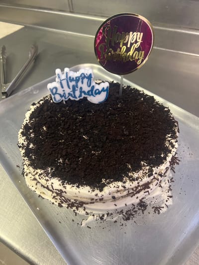 The image displays a round cake placed on a shiny, metallic surface, likely a tray or counter. The cake is predominantly covered on its top surface with a generous layer of dark brown crumbs, resembling crushed cookies (possibly Oreos). Around the perimeter of the cake, a thick layer of white frosting is visible, with dark brown sprinkles scattered across it and trailing off onto the metallic surface.
Two "Happy Birthday" decorations are inserted into the cake. To the left side of the cake, there is a round cake topper with a dark purple or maroon background featuring a marble-like pattern. Gold, metallic-looking script text spelling "Happy Birthday" is prominently displayed on this topper. Below and slightly to the right of this topper, a white, irregularly shaped candle or decorative figure is present, also bearing the words "Happy Birthday" in a glittery blue script.
The metallic surface beneath the cake reflects some light and shows minor scratches or marks. The overall impression is of a prepared birthday cake awaiting celebration. The image appears to be taken from a slightly elevated angle looking down at the cake, and it is rotated counter-clockwise, showing the metallic surface running vertically on the left and right sides of the cake.