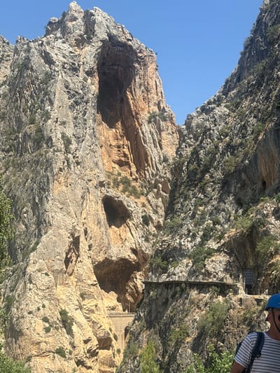The image captures a dramatic view of the Caminito del Rey, a walkway built into the side of a steep gorge in Ardales, Spain. Towering, rugged rock faces dominate the scene, with a large cave opening visible high up on the left. A narrow, suspended pathway, the Caminito del Rey itself, is affixed to the cliff face, appearing to wind its way through the gorge. In the foreground, a person wearing a blue hard hat and a striped shirt is partially visible, facing away from the camera, suggesting they are a visitor or hiker. The environment is characterized by steep, rocky terrain with sparse vegetation clinging to the slopes, under a clear blue sky. The scale of the rock formations and the precariousness of the pathway highlight the natural grandeur and the engineering feat of the walkway.