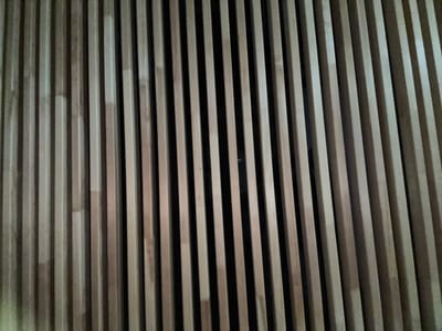 The image shows a close-up, top-down view of a wall made of vertical wooden slats. The slats are light brown and appear to be made of bamboo. There are dark spaces between the slats, suggesting a dark background or a deep recess behind the wall. The lighting is dim, casting shadows that emphasize the texture and verticality of the wood. There are no people or other identifiable objects in the image. The setting is indeterminate but could be an interior decorative wall or a privacy screen. The image does not contain information about the city of Shebarshino, Russia.