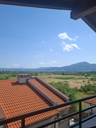 The image shows a view from a balcony overlooking a rural landscape. In the foreground, there is a terracotta tiled roof with a chimney. Beyond the roof, a field stretches out, dotted with trees and a few white animals, possibly cows. In the distance, rolling hills and mountains are visible under a clear blue sky with scattered white clouds. The location is described as the city of Ognyanovo, Bulgaria.