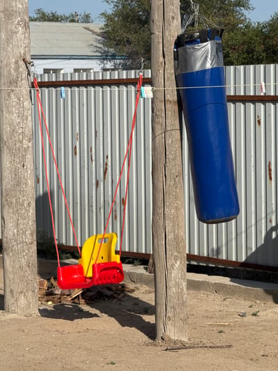 This image shows an outdoor scene in Аққыстау ауылдық округі, Kazakhstan. A large, blue punching bag is suspended horizontally from a wooden beam. It is attached with chains and silver duct tape. Below the punching bag, a yellow and red child's swing seat hangs from red ropes, also supported by the wooden beam. The background consists of a corrugated metal fence and some greenery. The ground appears to be dirt and gravel. The lighting suggests it is daytime, with strong shadows indicating a sunny day. There are a few clothespins visible on the fence, suggesting laundry may have been hung there. The overall impression is of a simple, utilitarian outdoor setup, possibly in a rural or backyard setting. No people are visible. There is no discernible text in the image, apart from what might be embossed on the punching bag, which is not legible.