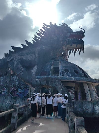 The scene is set outdoors in Kim Long Ward, Vietnam, beneath a partly cloudy sky with bright sunlight. The central subject is a large, derelict concrete dragon sculpture, intricately detailed and appearing weathered, with dark, discolored surfaces and visible peeling paint. The dragon is depicted in an aggressive stance, its head turned right with an open mouth revealing teeth and a curled tongue or tendril, and its body wraps around a domed, multi-level building. This underlying structure is also in disrepair, featuring visible graffiti in various colors and broken or empty window openings.
In the foreground, a group of approximately 10-12 individuals, predominantly women, stand on a concrete pathway or bridge equipped with railings. They are casually dressed, mostly in white or light-colored tops and dark bottoms, with many wearing white or light-colored hats. The individuals are positioned facing the dragon structure, appearing to observe it. Some are standing casually, while one person on the right is holding a device, possibly a phone, upwards as if taking a photograph. The strong sunlight from the upper center backlights the dragon, creating a silhouette effect and lens flare.