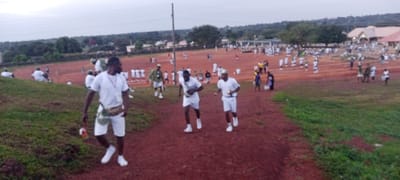 An outdoor scene in Peyer, Nigeria, depicts numerous individuals engaged in organized activity on a large red earth field. In the immediate foreground, a Black male walks towards the viewer on a red dirt path, wearing a white t-shirt, white shorts, white sneakers, and a tan fanny pack across his chest. He holds a small, colorful, striped object in his right hand. Slightly further back on the same path, two other Black males jog away from the viewer, also dressed in white t-shirts, white shorts, and white sneakers, each appearing to hold a small object.
The expansive mid-ground and background feature a red dirt field occupied by an estimated 100+ individuals, predominantly Black males, many dressed in white t-shirts and white shorts, forming various groups and lines, indicative of drills or physical training. Some individuals are observed in olive green or camouflage attire. A grassy embankment defines the left side of the foreground and parts of the mid-ground.
The distant background includes several buildings with light-colored roofs, scattered green trees, and residential structures, all under an overcast or uniformly lit sky. Utility poles are visible across the field.