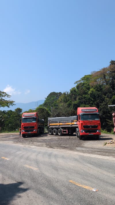 The image captures two red trucks parked on an asphalt road, partially obscured by trees. The scene is set in Lâm Sơn, Vietnam, as indicated by the "CTY CỔ PHẦN KHOÁNG SẢN & VLXD LÀM ĐỒNG" (joint stock company for minerals and construction materials), a business located in the area. The sky is a clear blue. A yellow line is painted on the road. The trucks are parked on the side of the road. The image is oriented sideways. A mountain is visible in the background.