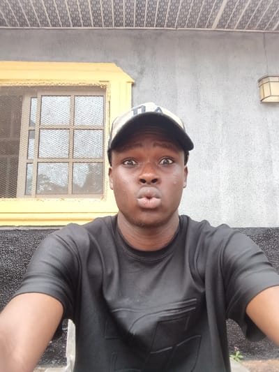 A male individual, appearing young and of African descent, is depicted in a forward-facing portrait. He wears a black t-shirt with a subtle, dark geometric pattern and a camouflage-patterned baseball cap with a dark emblem, worn brim-forward. His eyes are wide open, and his lips are pursed into a "fish-face" expression, directed towards the camera. The background consists of a building exterior. The upper wall section is light grey, possibly stucco or concrete. A yellow-framed window, fitted with a white security grille and multiple glass panes, is visible to the left. Below the window, the wall transitions to a darker, textured grey-black finish. An octagonal, light-colored wall sconce is mounted on the upper grey wall to the right. Above the wall, a patterned ceiling or soffit, possibly an overhang, with a grid design is visible. Small plant growth is present at the bottom right corner of the frame. The lighting is even, indicating an outdoor daytime setting. The specific location in Itu, Nigeria, cannot be determined from the visual information provided within the image.