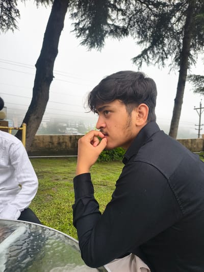 Two individuals are seated at a table with a glass top in an outdoor setting, likely in Khaniara, India, given the context. The person on the right, in the foreground, is a young male wearing a black collared shirt and black pants. He is captured in profile, looking to his left, with his right hand raised to his mouth, appearing to be touching his mustache or chin. His dark hair is styled with some volume. To his left, partially visible, is another person wearing a white collared shirt and black pants. They are seated and facing partially towards the foreground. The environment includes lush green grass, trees with dark bark and foliage, and a blurred background suggesting buildings and a misty, possibly overcast sky. A low stone wall is visible behind the seated individuals, and a portion of a gold-colored railing or fixture is also present. The overall lighting is diffused, indicating an overcast day or a shaded location.