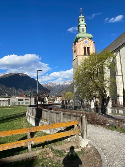 The image showcases a combination of architectural and natural elements in what appears to be Sterzing-Vipiteno, Italy. In the foreground, there's wooden construction scaffolding with a partial view of a grassy area and a building with a modern facade, featuring rows of windows. Beyond this foreground scene, a mountain range with some snow-covered peaks is visible under a bright blue sky with scattered clouds.
The top portion of the image features the steeple of a church, painted in pastel colors, including green and pink. A tree partially obscures the church's exterior, with branches and leaves visible. A section of a stone building and a paved area is seen adjacent to the church.