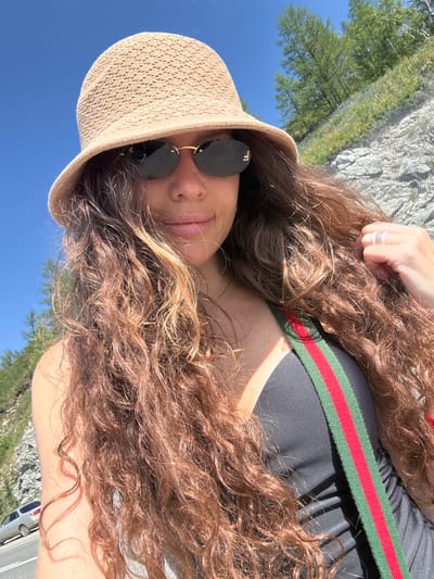 A woman wearing a straw bucket hat and dark sunglasses is captured in a selfie with a bright blue sky and a wooded, rocky hillside in the background. She has long, curly brown hair that partially obscures her face. A green and red striped strap is visible across her chest, and she is wearing a black garment. A ring is visible on her right hand. The scene appears to be outdoors, possibly in a mountainous or hilly area. No specific location in Khabarovka, Russia, can be determined from the visual information.