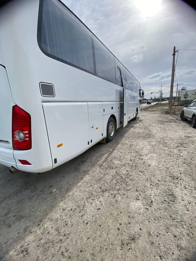 A large, white, modern tour bus is parked on a rough, unpaved gravel road, casting a shadow to its right. The image is taken from a low angle, looking upwards and along the side of the bus towards the front. The sky is bright and overcast, with the sun prominently visible on the left side, indicating daytime. Utility poles with wires run along the roadside in the distance.

The bus features large, dark-tinted windows running along its side and a distinctive large rear window. Multiple compartments are visible on the side of the bus, along with a rear taillight assembly and several vents. The gravel ground is light grey, composed of small stones and dirt.

In the background, beyond the bus and utility poles, there are distant buildings, other parked vehicles (including a white car), and a large billboard displaying Cyrillic text, including "Б" and "СКВ", and a visible Russian flag.

Visible text on the side of the bus, in blue lettering, appears to read "BELOYAR". At the very rear of the bus, near the taillight, the model name "GRAND BIRD" is discernible. The scene suggests a transit area or parking lot on the outskirts of an urban or semi-urban environment in Bol'shoe Teshkovo, Russia. No people are visible.