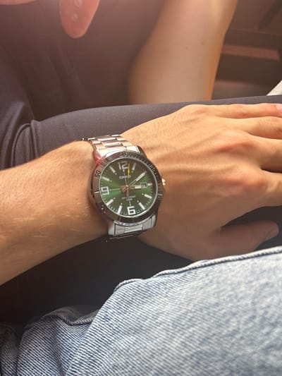 The image shows a close-up of a person's wrist wearing a silver Casio watch with a green dial. The watch has a metal band and shows the time as approximately 12:05. The background is blurred, but appears to show dark fabric and denim. The location of Ivlevo, Russia, is not visible or implied in the image.