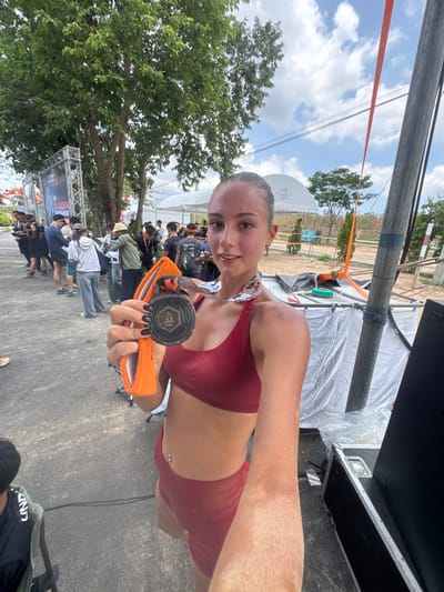 A woman is holding a medal in her right hand, looking into the camera and smiling. She has a maroon sports bra and shorts and the medal is silver with an orange band. Behind her, other people are standing in line. There is a tree and a banner that reads "Mudder Finisher". There is a white tent in the background. The location is บ้านสำนักตะแบก, Thailand.