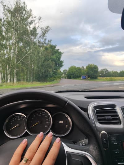 This is a photograph taken from inside a car, looking forward through the windshield. The dashboard is visible in the foreground, with the steering wheel on the left and the instrument cluster in the center. The speedometer shows a reading of 50 km/h, and the odometer shows 1856. A woman's hand with manicured nails rests on the steering wheel. Outside the car, a tree-lined road is visible, with a cloudy sky overhead. The location is identified as Kachalivka, Ukraine. A small blue structure is visible in the distance.