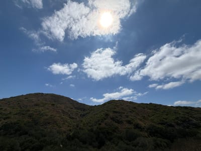 The original image is a landscape photograph taken on a sunny day in Laguna Beach, California, United States of America. The photo depicts a clear blue sky with scattered white clouds and a bright sun partially obscured by a cloud. In the foreground, there are two green, tree-covered hills. The hills are densely vegetated with a mix of dark green trees and lighter green shrubs. The sun is positioned in the upper center of the sky, casting a bright light and creating a slight glare. There are no people or animals visible in the image. The overall scene conveys a natural, outdoor environment with a focus on the sky and the rolling hills.