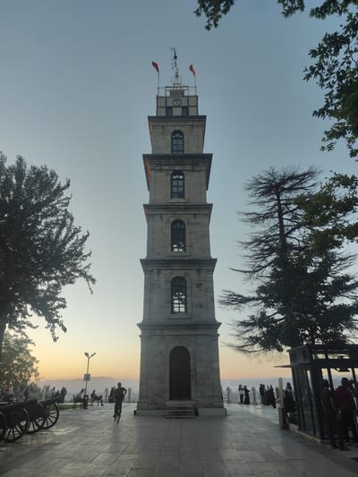 This media file captures a view of the Tophane Clock Tower in Osmangazi, Turkey, during what appears to be sunset or dusk. The tall, stone clock tower is the dominant subject, with its classical architecture featuring arched windows and a prominent clock face near the top. A red flag flies from the tower's peak, alongside some metallic structures.

The scene is set in an open public area, paved with large stone tiles. In the foreground, to the left, are several historical cannons lined up, suggesting a historical site or park. People are scattered throughout the scene, enjoying the late afternoon or early evening atmosphere. Some individuals are seated on benches, while others are walking or cycling. In the mid-ground, a person is riding a bicycle, creating a sense of movement. Further back, a group of people are gathered near a railing overlooking a distant cityscape and hills, silhouetted against the soft, warm light of the setting sun. Trees frame the tower and the surrounding area, with lush green foliage on the left and a more barren, spiky tree on the right, adding depth to the composition.

The sky transitions from a pale blue at the top to a warm gradient of orange and yellow near the horizon, indicating the time of day. The overall mood is peaceful and serene, with people relaxing and appreciating the view. There are no visible signs or text within the frame.