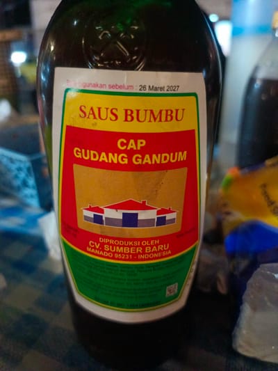 The image shows a close-up of a bottle of "SAUS BUMBU CAP GUDANG GANDUM" sauce. The label is mostly visible, displaying text and graphics related to the product. At the top of the label, it says "Baik digunakan sebelum: 26 Maret 2027". Below this, in large yellow lettering, is "SAUS BUMBU", and then in red lettering "CAP GUDANG GANDUM". There is an illustration of three stylized houses with red roofs and blue and white walls. Further down, it states "DIPRODUKSI OLEH CV. SUMBER BARU MANADO 95231 - INDONESIA". The bottle appears to be dark green glass. The background is blurred and shows hints of other bottles and objects, suggesting an indoor environment like a store or pantry. There are no people or any identifiable landmarks that would suggest the specific location in the city of Luwoo, Indonesia.