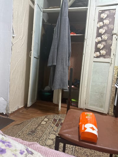 The image depicts an indoor scene, possibly a cluttered room or hallway, with a prominent wardrobe or closet on the right side of the frame. The wardrobe doors are partially open, revealing shelves within, one of which holds clothing and a green plastic container. A gray towel or garment hangs from the left side of the wardrobe. The flooring appears to be wooden planks covered in a patterned rug. In the foreground, a brown cushioned surface, possibly a chair or stool, holds an orange cardboard box from "storkebab" with text that appears to be in Cyrillic script. The walls show signs of wear, with peeling wallpaper on the left. The overall impression is one of a lived-in, somewhat disheveled domestic space. There are no people visible in the image, and no specific landmarks or contextual clues suggest a location within the city of Corlăteni, Moldova.