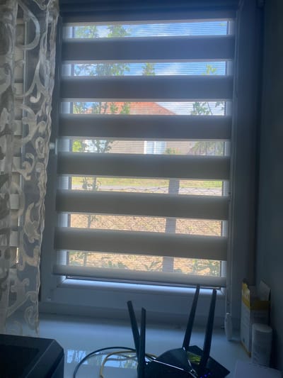 The image captures a view from a window, looking outward through a dual-layer roller blind. The blind consists of alternating horizontal opaque and sheer fabric panels, allowing adjustable light and visibility. Outside, a residential area is visible, with the roof of a house with terracotta tiles and green foliage from trees interspersed with glimpses of the sky. The window is situated on a windowsill, where several objects are placed. These include a black computer tower on the left, a wireless router with multiple antennas positioned towards the right, and a yellow and white cardboard box with text, along with a white cylindrical object, likely a container, to its right. A sheer curtain with a decorative pattern is drawn to the left of the window. The overall impression is an indoor scene in a residential setting, with a view of a typical Ukrainian suburban or town environment. The specific location in Khmilnyk, Ukraine, is not determinable from the visual information provided.