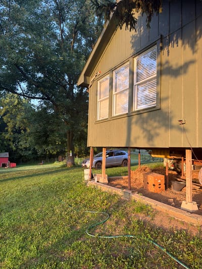 The image depicts a side view of a light green, wooden house raised on stilts, located in Holt, United States. The house has two windows with blinds. Underneath the house, a silver SUV is parked, and there is evidence of construction or storage materials, including hay bales and wooden beams. A cardboard box with ventilation holes sits on the hay. A green garden hose snakes across the grassy lawn in the foreground, which has scattered white flowers. In the background, a large, leafy tree stands prominently, and to the left, a small red shed with a red object next to it is visible. The scene is bathed in the warm light of what appears to be either sunrise or sunset, casting long shadows.