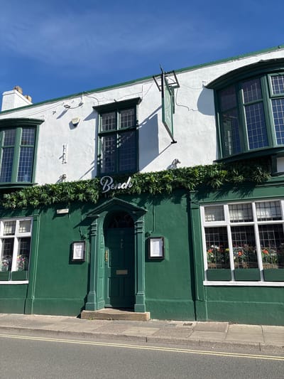 The image shows the exterior of a building in Nantwich, United Kingdom, which appears to be a restaurant or bar called "Bench." The building has a white upper story with green trim and windows, and a dark green lower story with a prominent arched doorway. A green sign with the name "Bench" is mounted above the doorway. The building is decorated with artificial greenery along the facade. There are several windows with plants visible inside. A street with double yellow lines runs in front of the building. The sky is clear and blue.