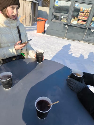 The scene depicts an outdoor gathering during daylight in a snowy environment in Prybuzke, Ukraine. A round, dark grey table occupies the foreground and midground, covered in sunlight and shadows. Four black, corrugated paper cups containing hot beverages are placed on the table; three hold a dark liquid, emitting steam, and one contains a lighter liquid with a lemon slice. A wooden stir stick is present in one of the dark liquid cups.
On the left side of the table, a young female, wearing a brown knitted beanie and a cream-colored puffer jacket with a fur-lined collar, holds a smartphone in her left hand and one of the black paper cups in her right. A black shoulder bag with a gold clasp hangs from her left shoulder. Her mouth is slightly ajar.
On the right side of the table, a second individual's gloved hands, clad in dark grey, are seen holding another black paper cup. The hands are positioned as if warming them on the cup.
The ground surrounding the table is covered in an unbroken layer of white snow, with multiple elongated shadows of people cast upon it, suggesting the presence of at least two individuals, possibly more. In the background, an orange wheeled garbage bin with a black liner stands on the snow, adjacent to a dark grey commercial kiosk or stand with windows displaying packaged food items and drinks. Behind the bin, a light-colored, tent-like structure or shed is visible, with bare trees and a fence beyond it. The sky is clear, indicating bright sun conditions.
