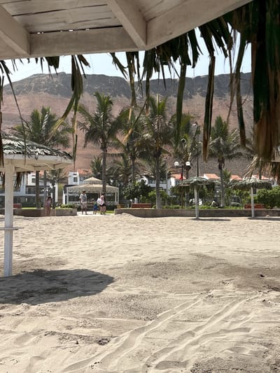 The image shows a sunny beach scene in San Antonio, Peru, with a view of palm trees, buildings, and a mountainous landscape. In the foreground, a sandy beach stretches out, with tire tracks and footprints visible in the sand. A portion of a white thatched umbrella is visible on the left side of the frame, casting a shadow on the sand. In the mid-ground, a few people are walking on the beach, including a family with children. Behind them, there are several buildings with white walls and red tiled roofs, suggesting a resort or hotel complex. Palm trees are scattered throughout the area, and in the background, a large, dry, reddish-brown mountain rises against a clear blue sky. The overall impression is one of a tropical or subtropical coastal environment.