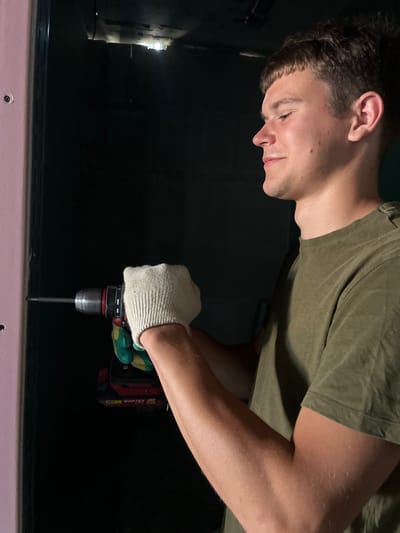 A young man with short brown hair, wearing a green t-shirt and white work gloves, is shown from the chest up, in profile, to the right of the frame. He is holding a red and black cordless drill with a silver chuck and a long metal bit, which is engaged with a pink surface on the left side of the image. The drill has "18V WORTEX" visible on it. The background is predominantly dark, with a faint light source visible in the upper center, suggesting an interior space. The overall scene depicts a do-it-yourself or construction activity within what appears to be a room in Аснежыцкі сельскі Савет, Belarus.