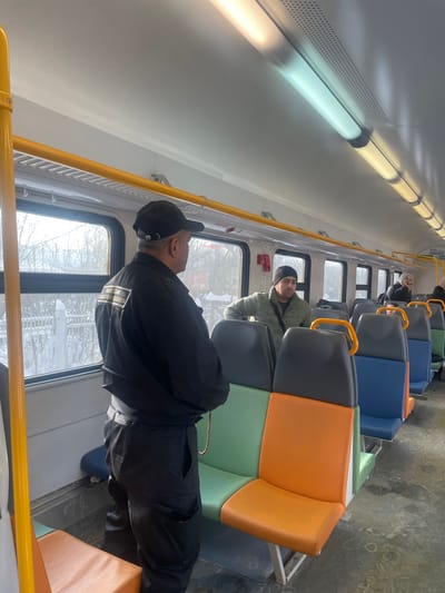 The image displays the interior of a commuter train car, specifically located in Fryazevo, Russia, during daylight hours. A man in a black uniform, including a black baseball cap and a jacket featuring a light-colored rectangular patch with Cyrillic text on the upper back, stands with his back partially to the viewer, facing another man. A string of reddish-brown beads hangs from the uniformed man's left side. The second man wears a dark beanie and an olive-green jacket, and is positioned further down the aisle, appearing to lean against a seat while looking towards the uniformed individual.
The train car features a white ceiling equipped with multiple fluorescent tube lights, some emitting a cool white light and others a warmer yellow light. Yellow grab rails are mounted above the windows, through which a snow-covered exterior landscape with trees and a white fence is visible. The seating consists of rows of multi-colored upholstered seats, featuring segments of green, orange, blue, and dark gray. A red emergency call button is mounted on the wall between two windows. Several other passengers are partially discernible further along the aisle in the background. The floor appears to be a dark, textured surface.