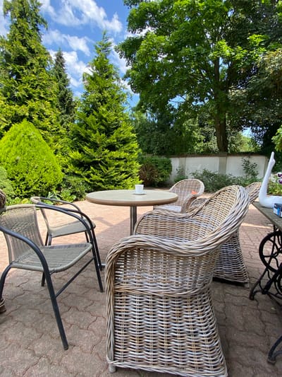 The image depicts a tranquil outdoor seating area in Wielka Wieś, Poland. The foreground features two chairs, one made of woven rattan with a curved back and another with a metal frame and woven seat and back. A small, round table with a light-colored surface stands between them. In the background, a large rattan chair with a distinctive curved back is prominent. Beyond the seating area, lush green trees and shrubs create a natural backdrop, with a clear blue sky and scattered white clouds visible overhead. A white wall can be seen behind the rattan chair. The scene suggests a peaceful setting, possibly a garden or patio, designed for relaxation. The lighting indicates it is daytime, with sunlight casting shadows on the paved ground. There is no discernible activity or event taking place, and no visible text.